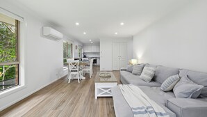 A modern living room with a grey sofa, a dining table with chairs, and a kitchen in the background.