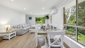A modern living room with a glass dining table, white furniture, and a large window overlooking greenery.