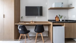 A modern kitchen with a flat-screen TV, a microwave, and a refrigerator. There are two black chairs and a wooden desk. - Zoom Santa Cecília by Viva - Prédio próximo à estação Marechal Deodoro e Fácil acesso ao Memorial da América Latina (São Paulo)