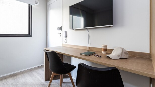 Un espacio de trabajo moderno con un escritorio de madera, dos sillas negras, un televisor montado y una ventana con persianas.