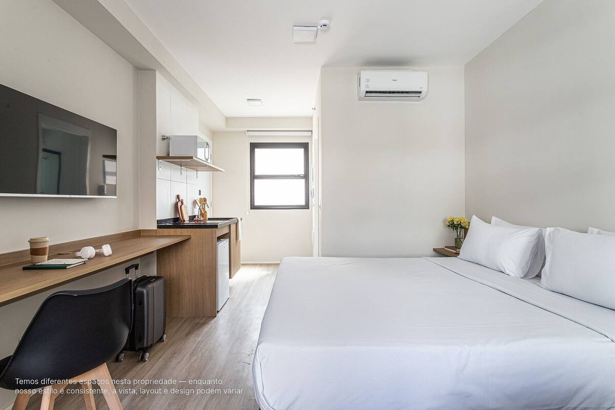 A modern hotel room with a large bed, a desk with a computer, a chair, and a suitcase.