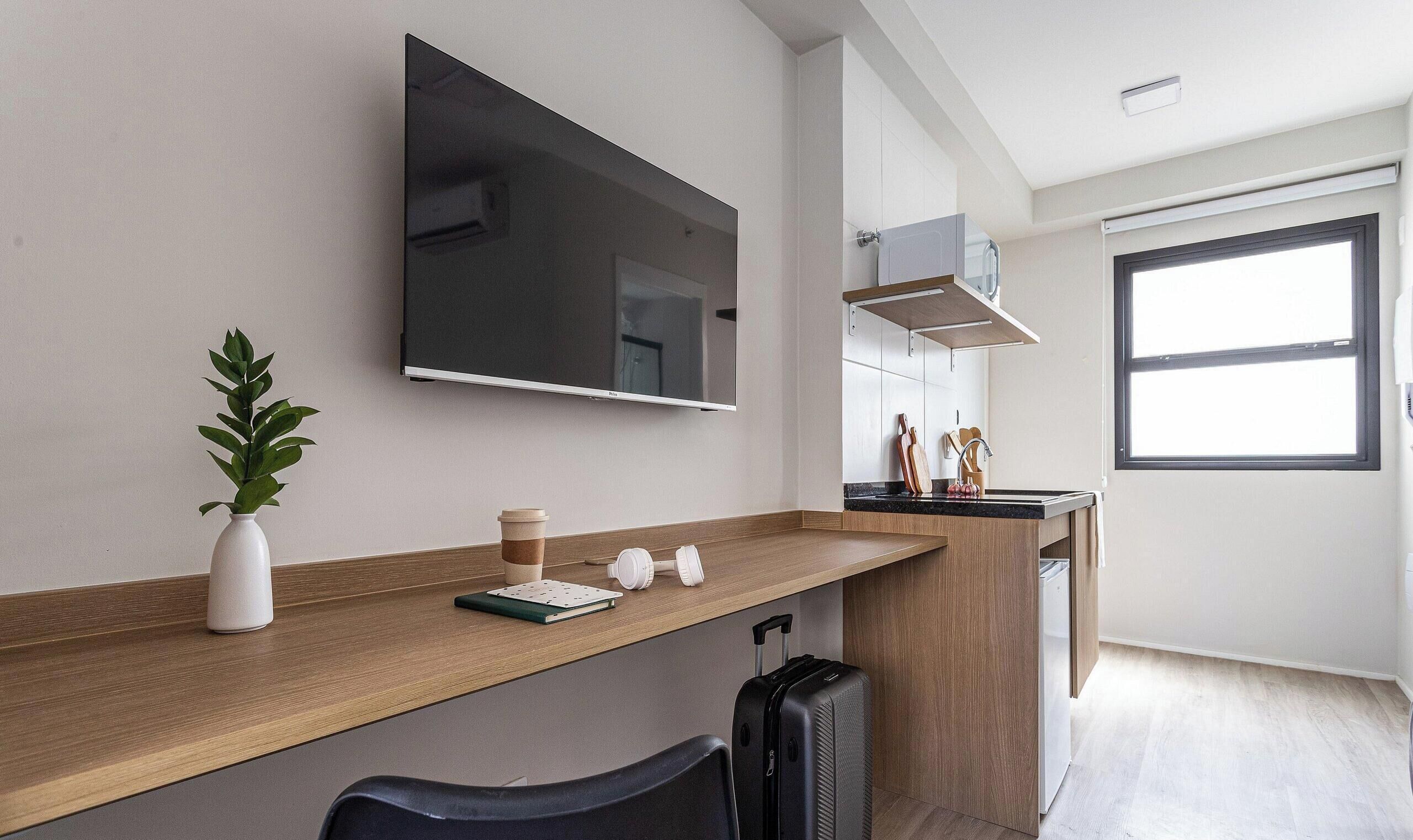 A modern hotel room with a wooden desk, a flat-screen TV, a potted plant, a coffee cup, a notebook, and a suitcase.