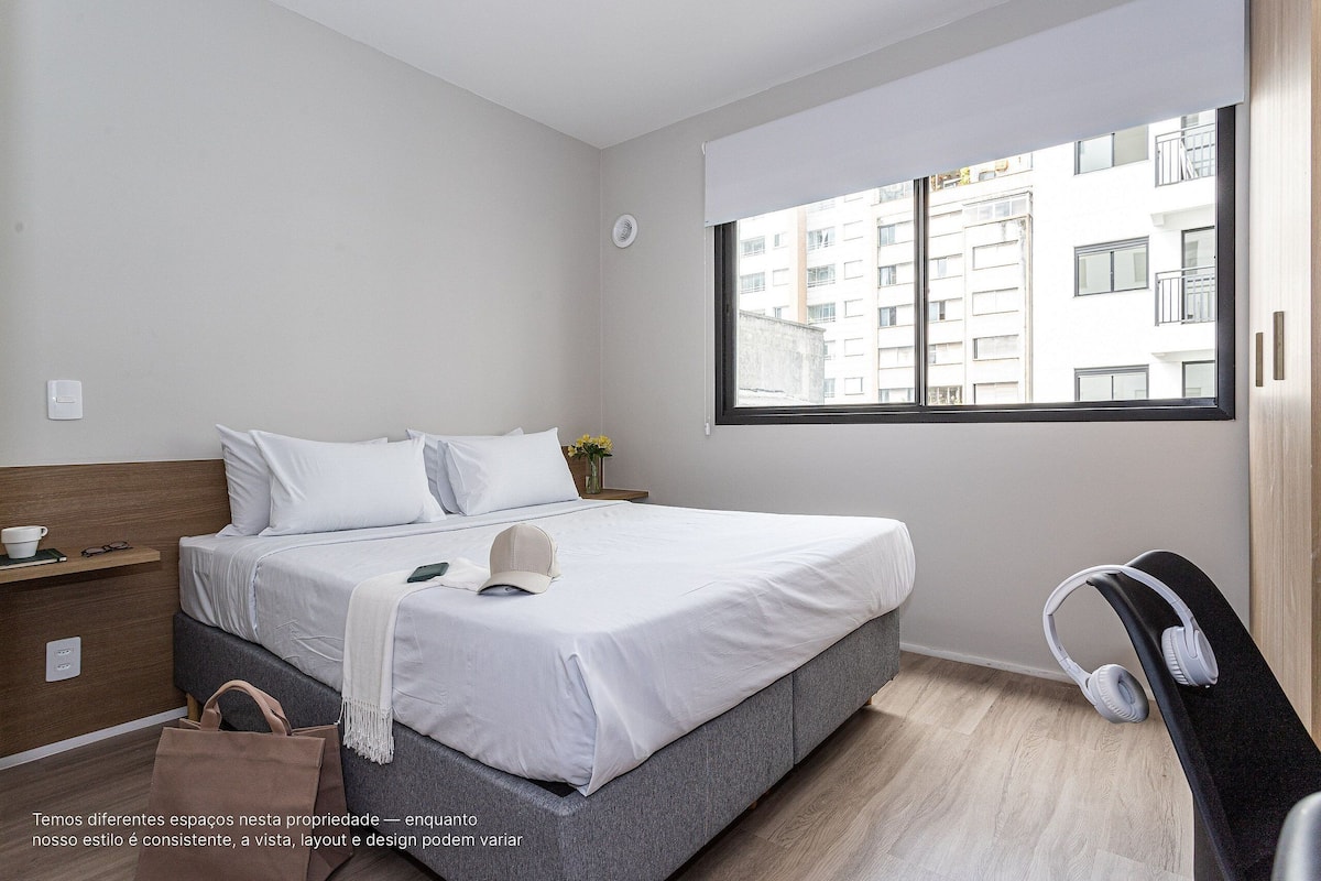 A modern hotel room with a large bed, a window with a city view, a bedside table, and a chair with headphones.