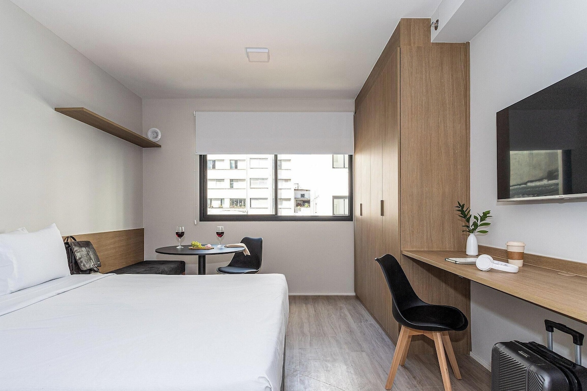 A hotel room with a bed, a desk, a chair, a small table, a window, and a TV.