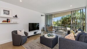 A modern living room with a flat-screen TV, grey sofas, a coffee table, and a balcony with outdoor furniture. - 2 Bedroom Garden & Pool View (Casuarina)