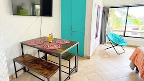 A small room with a wooden table, a bench, a teal-colored wardrobe, a TV mounted on the wall, and a window with a view of palm trees.