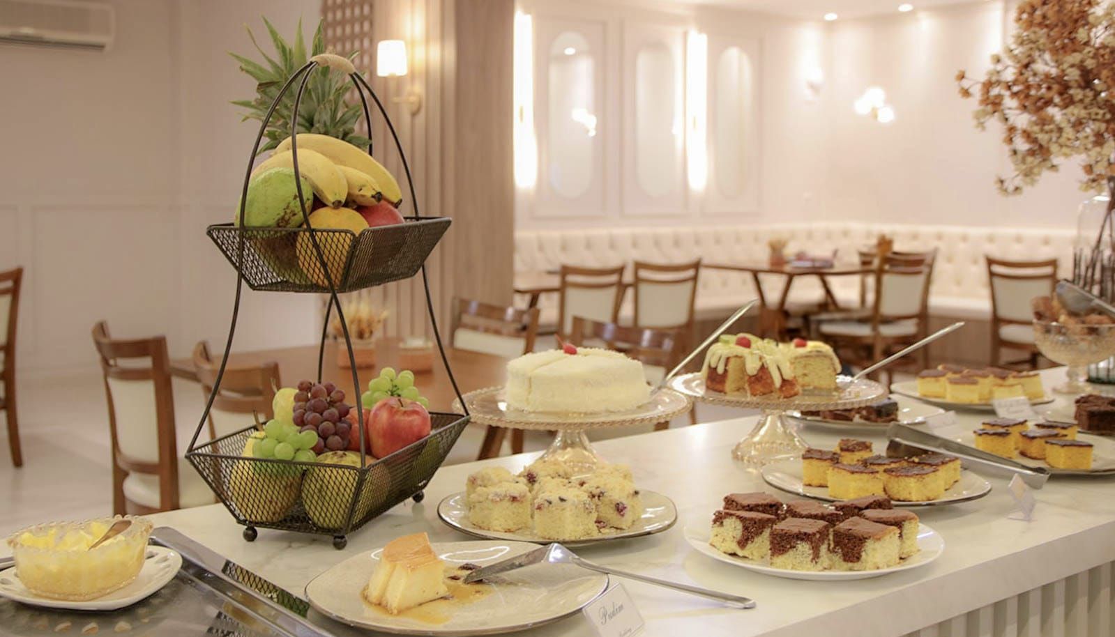 Uma mesa de buffet com diversos doces e frutas, incluindo bolos, tortas e uma cesta de frutas.