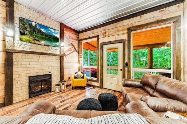 Una sala de estar acogedora con chimenea, sofá de cuero y un gran ventanal con vistas al bosque.