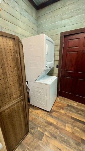 A compact laundry room with a stacked washer and dryer, a wooden door, and a wooden floor.