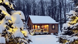 Une cabane en rondins dotée d’un porche décoré de guirlandes lumineuses et de festons, entourée d’arbres couverts de neige.