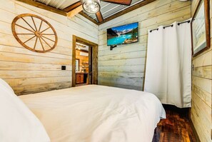 A wooden cabin room with a large bed, a mounted TV displaying a mountain scene, and a rustic wooden wheel on the wall.