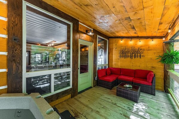 Una terraza de madera con un sofá rojo y una pequeña mesa, un ventanal de vidrio que da al exterior, y una pared con un diseño decorativo de árbol.