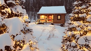 Una cabaña de troncos con techo cubierto de nieve, rodeada de árboles cargados de nieve y una terraza de madera al frente.