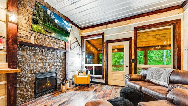 A cozy cabin interior with a stone fireplace, leather sofas, and a large window offering a view of the forest. - EvergreenGrove-2 cabins/4BR (Oakland)