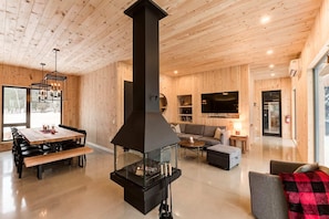 Ein modernes Interieur mit einem zentralen schwarzen Kamin, Holzwänden, einem Essbereich mit Tisch und Stühlen, einer Couch und einem an der Wand montierten Fernseher.