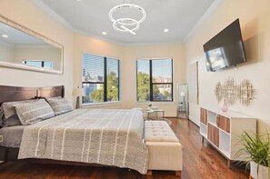 A modern bedroom with a large bed, a TV mounted on the wall, and a window with blinds.