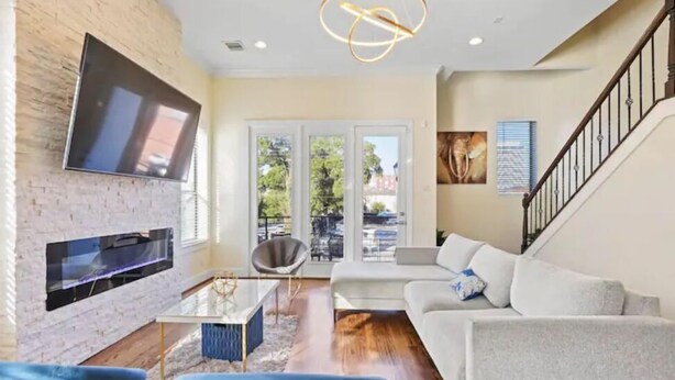 A modern living room with a flat-screen TV mounted on a brick wall, a sofa, a coffee table, and a staircase. - Luxury Midtown Gem : Amazing Rooftop Views (Houston)