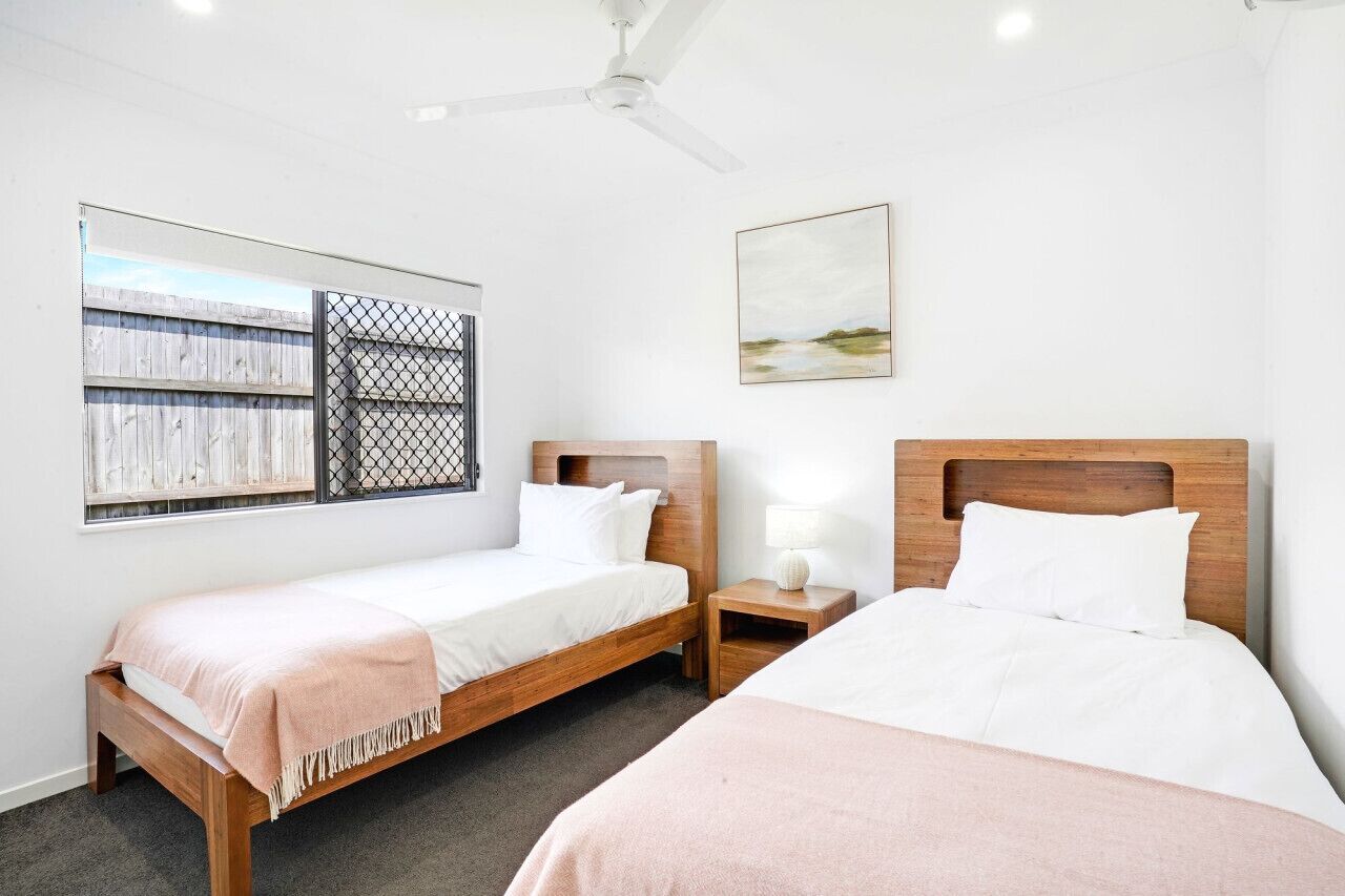 A hotel room with two wooden beds, a ceiling fan, a window with a view of a fence, and a painting on the wall.