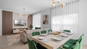 Un espace de salle à manger moderne avec une longue table dressée pour six personnes, des chaises à l’assise verte et un lustre doté de plusieurs points lumineux.