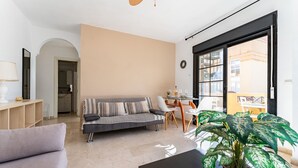 A living room with a sofa, a coffee table, and a potted plant. There is a dining area with a table and chairs.