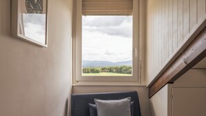 Una stanza con una finestra che si affaccia su un paesaggio verde, un divano grigio con cuscini e una scala in legno.