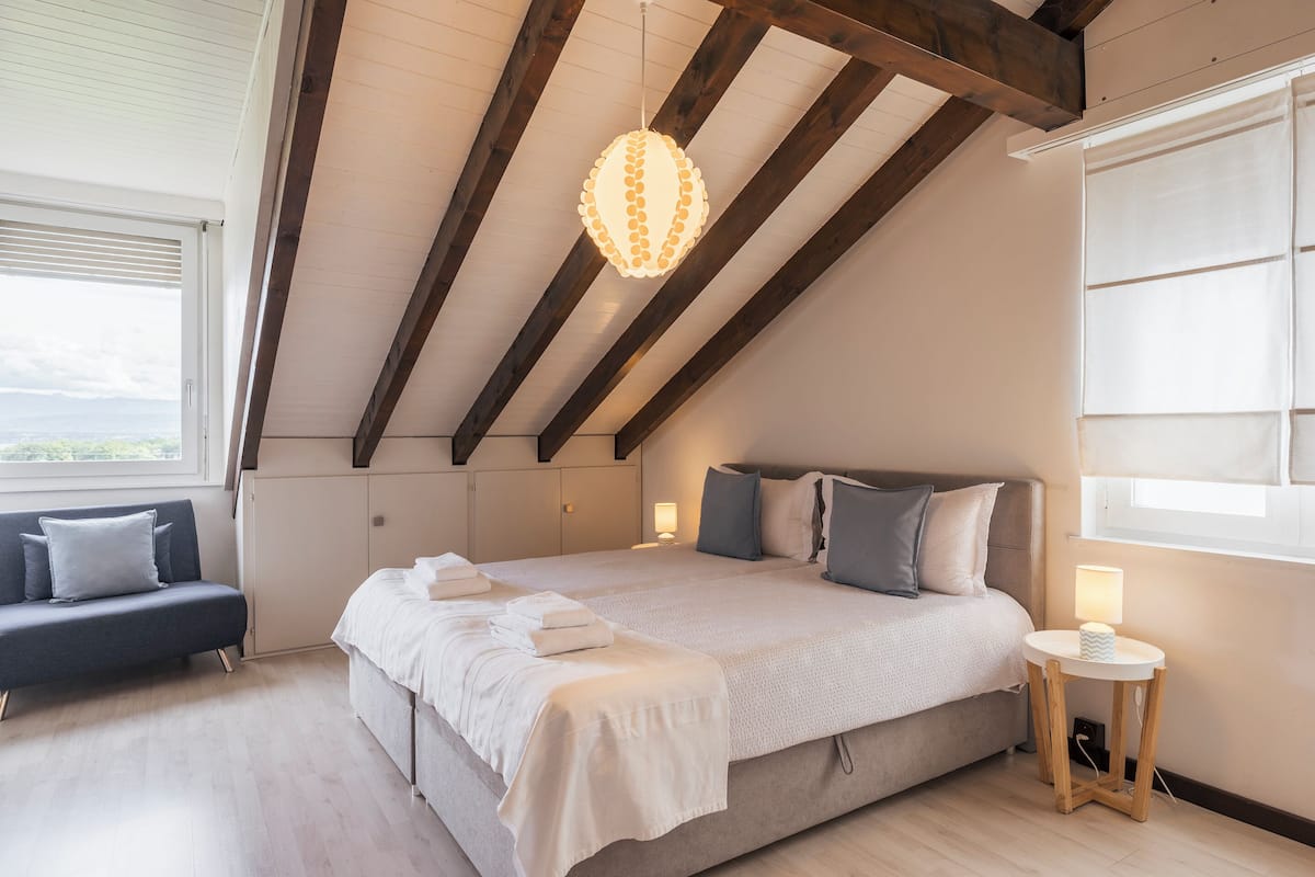 A bedroom with a large bed, a bedside table with a lamp, a bench, and a view of the outdoors through a window.