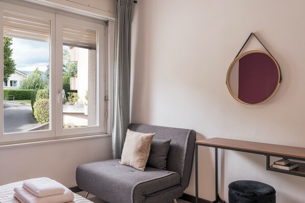 Et moderne værelse med en grå sofa, et lille bord, et rundt spejl og et vindue med udsigt til en gade og grønne områder.