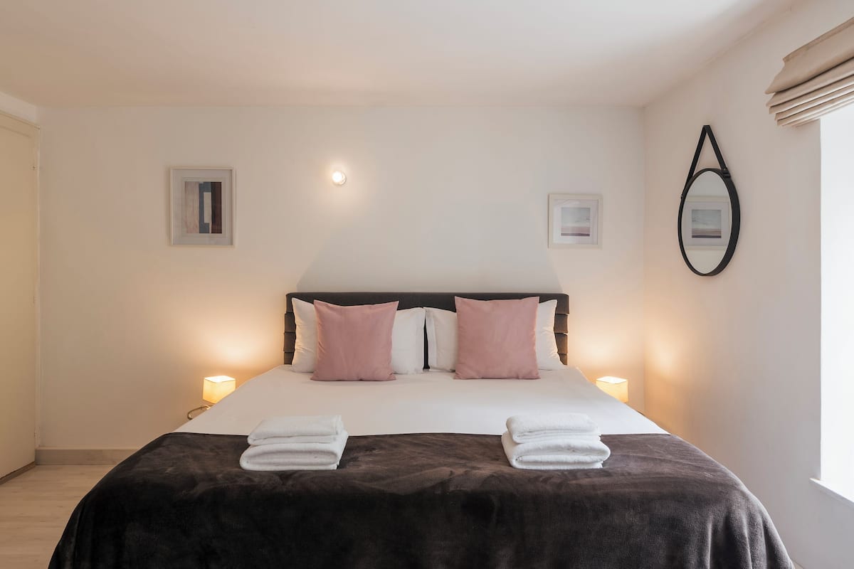 A neatly made bed with a dark brown cover, two white pillows, and two pink pillows. There are two bedside lamps on either side of the bed.