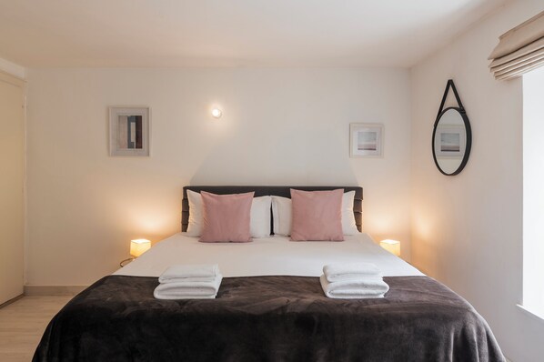 A neatly made bed with a dark brown cover, two white pillows, and two pink pillows. There are two bedside lamps on either side of the bed. - Amanthos Living Nyon (Self Check In) (Duillier)