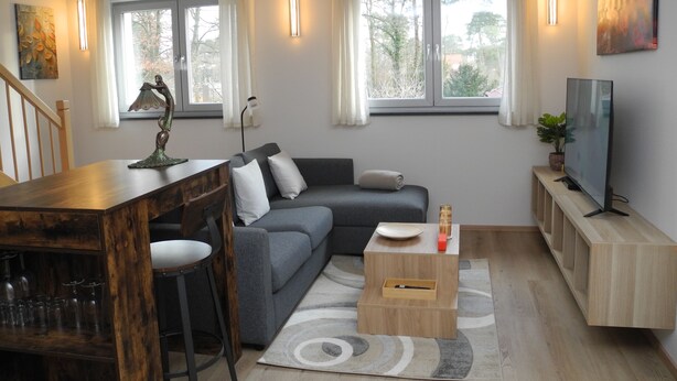 A modern living room with a grey sofa, wooden coffee table, and a TV on a wooden stand. There are two windows with curtains, a wooden dining table with chairs, and a staircase in the background.