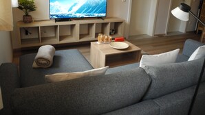 A living room with a grey sofa, a flat-screen TV on a wooden stand, a small table with glasses, and a potted plant.