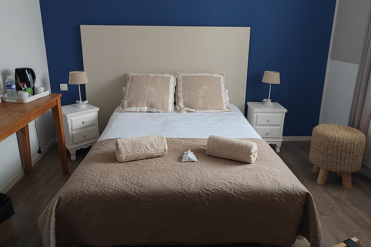 A hotel room with a large bed, two bedside tables, a nightstand, a wooden desk, a wicker ottoman, and a blue accent wall.