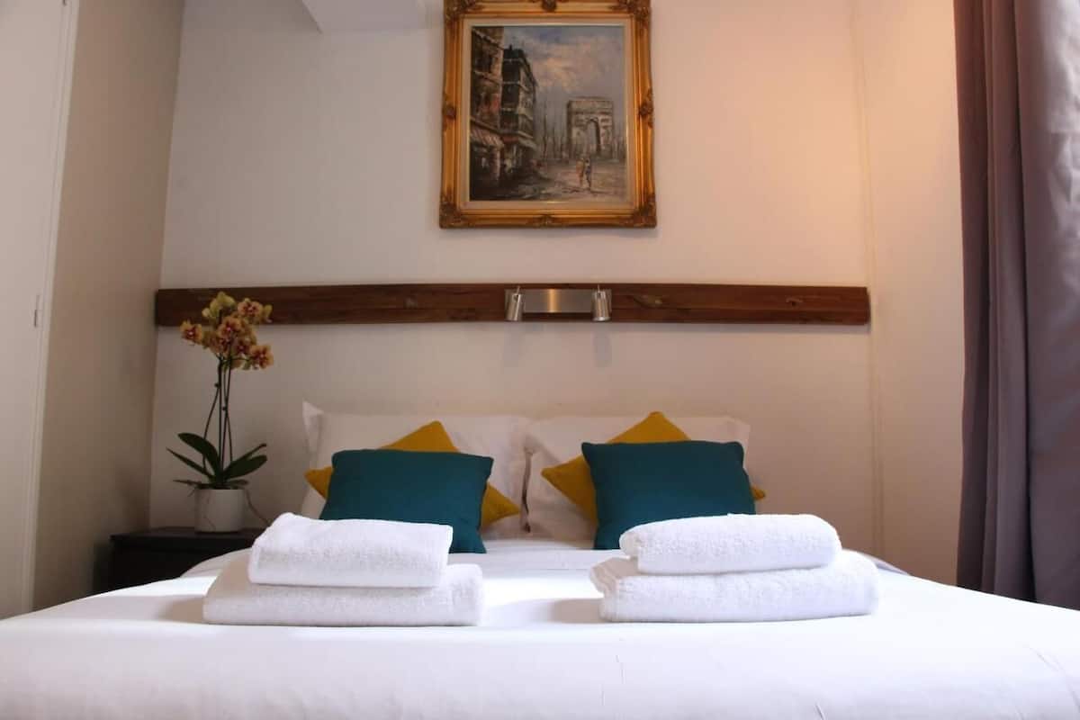 A neatly made bed with white linens and two towels, a painting of a historical building on the wall, and a potted plant on a bedside table.