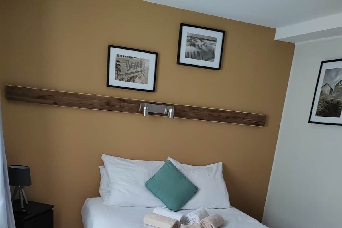 A neatly made bed with white linens, a green pillow, and rolled towels. There are two framed pictures on the wall above the bed.