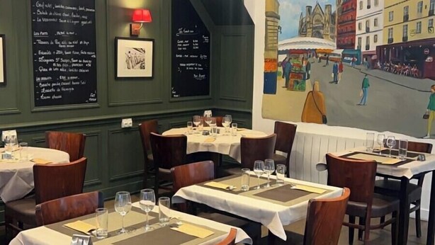 A restaurant interior with round tables, wooden chairs, and a mural on the wall depicting a street scene. - Hôtel Au Grand Duquesne  (Dieppe)