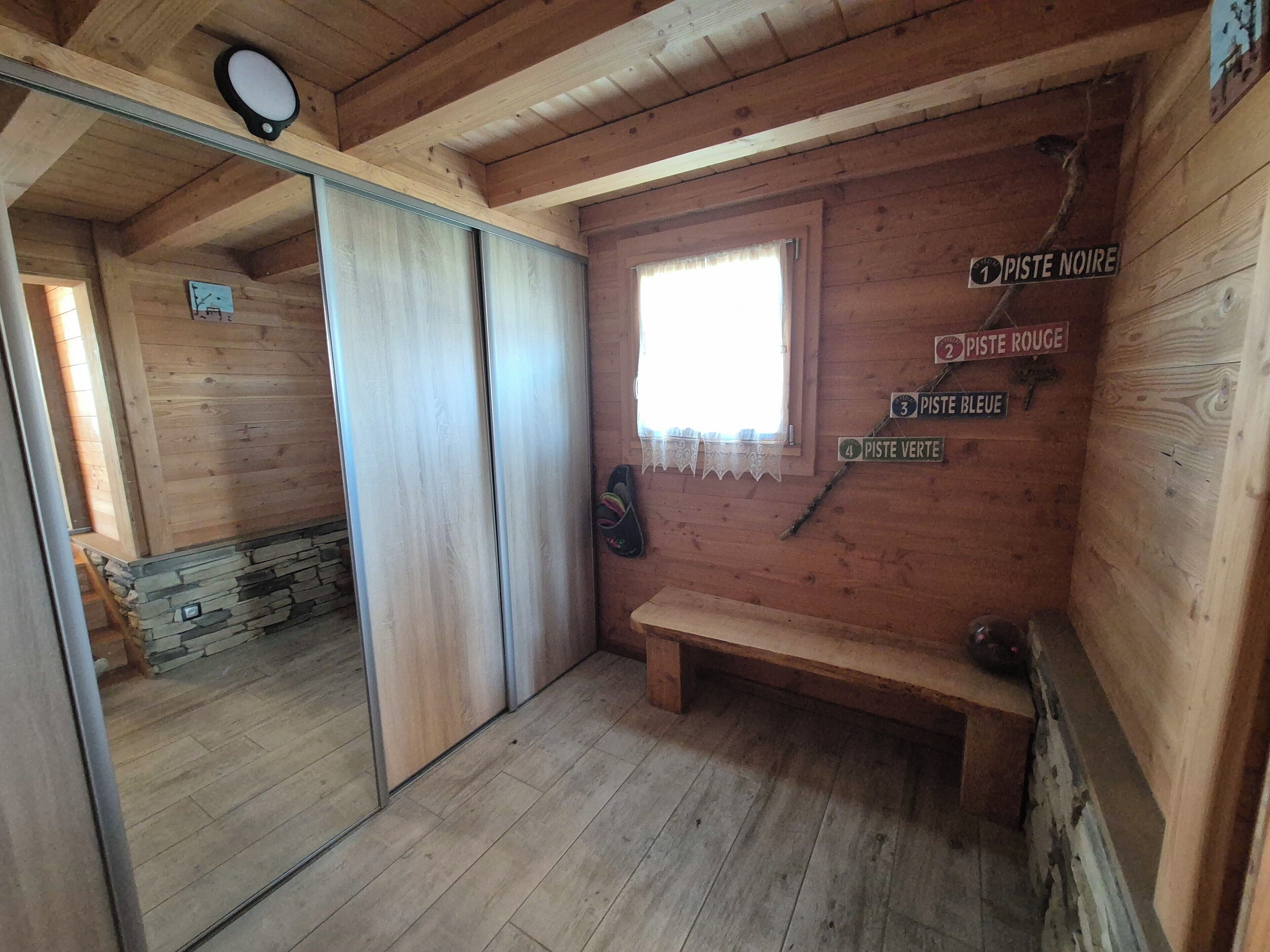 A wooden cabin with a stone wall, a wooden bench, and a window. There are signs indicating different rooms: Piste Noire, Piste Rouge, Piste Bleue, and Piste Verte.