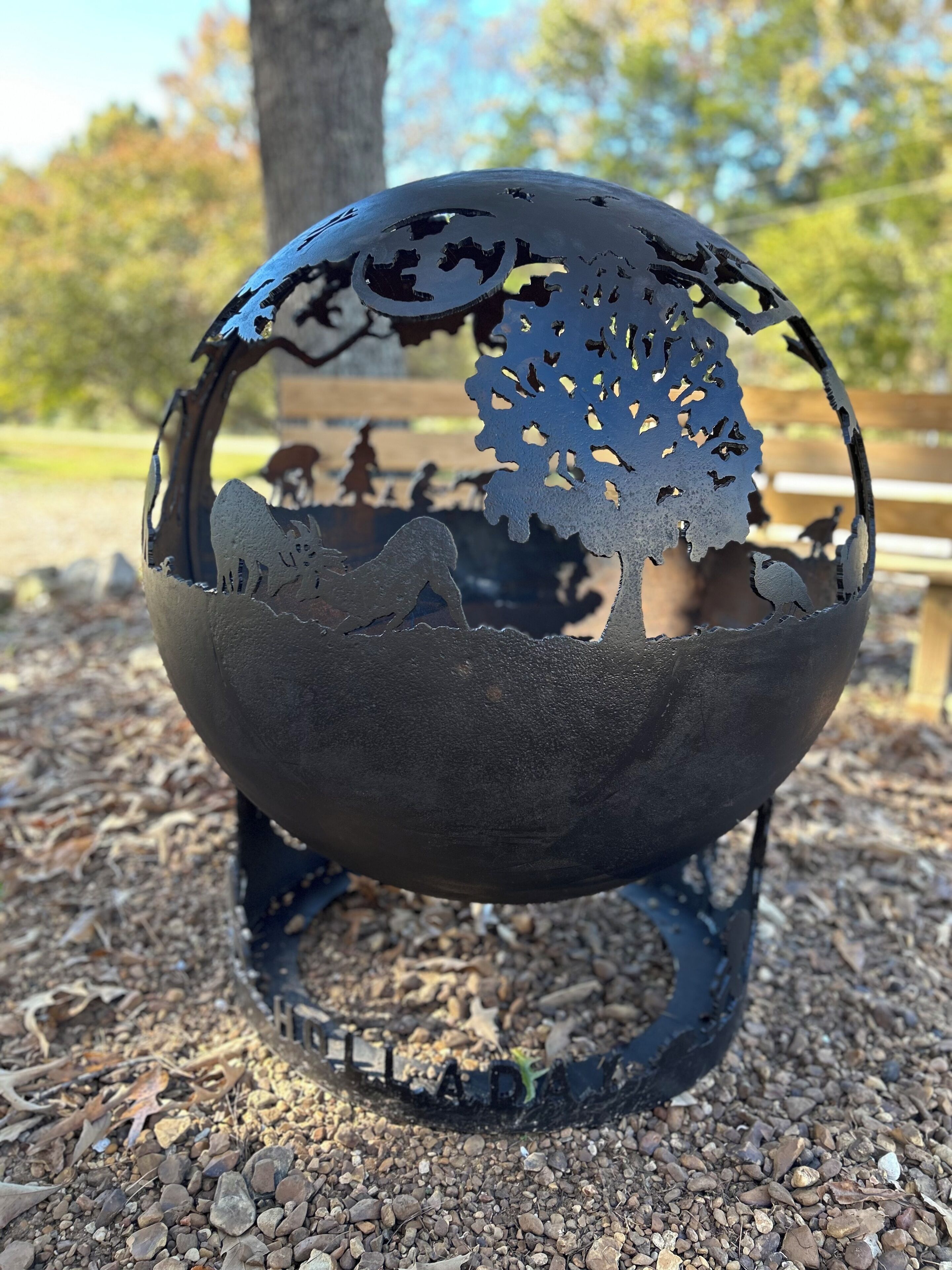 Eine Metallskulptur eines Baumes mit runder Basis, draußen auf einem Kiesuntergrund.