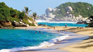 A beach with clear turquoise water, people swimming and walking on the sand, and large boulders in the background. - Taganga Tropical House (Santa Marta)