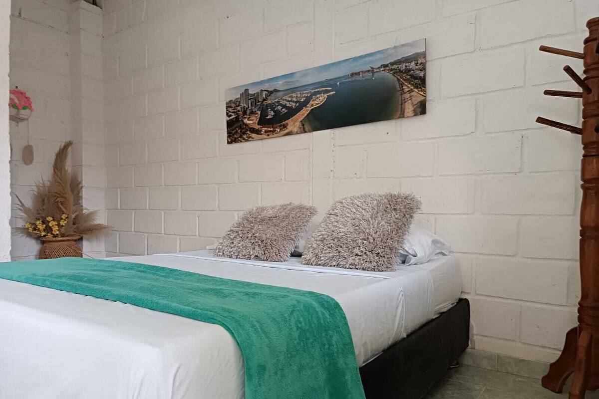 Un dormitorio con una cama, una manta verde, dos almohadas mullidas, un cuadro en la pared y una silla de madera.