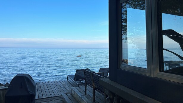 A wooden deck with a grill, a chair, and a table overlooking a calm body of water.