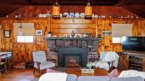 A cozy cabin interior with a stone fireplace, wooden walls, a dining area, and a television. - Charming 3-bedroom lakefront house with large yard and sandy beach (Colchester)