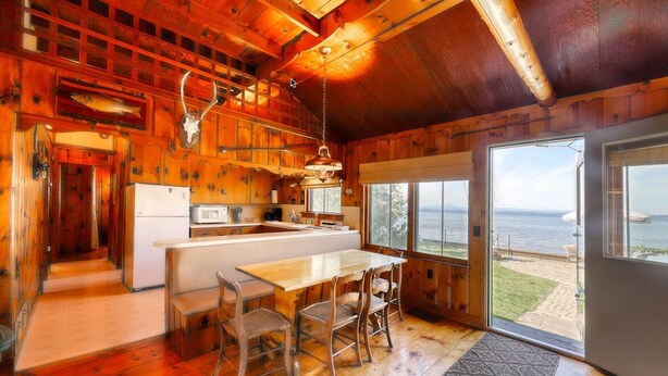 A wooden cabin interior with a kitchen, dining area, and large windows overlooking a beach. - Charming 3-bedroom lakefront house with large yard and sandy beach (Colchester)