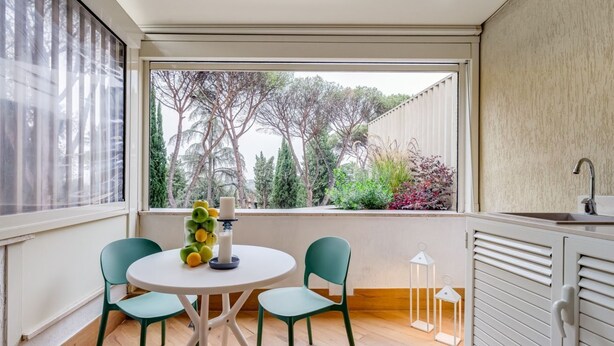 A compact room with a round table, two chairs, a window with a view of trees, and a kitchenette area. - BDC - Cassia Luxury Studio (Roma)