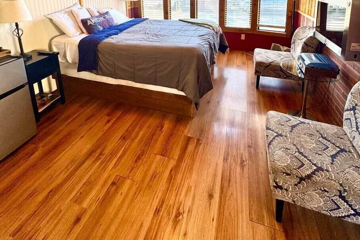 A cozy room with a wooden ceiling, a bed with a blue blanket, a desk with a lamp, and a chair.