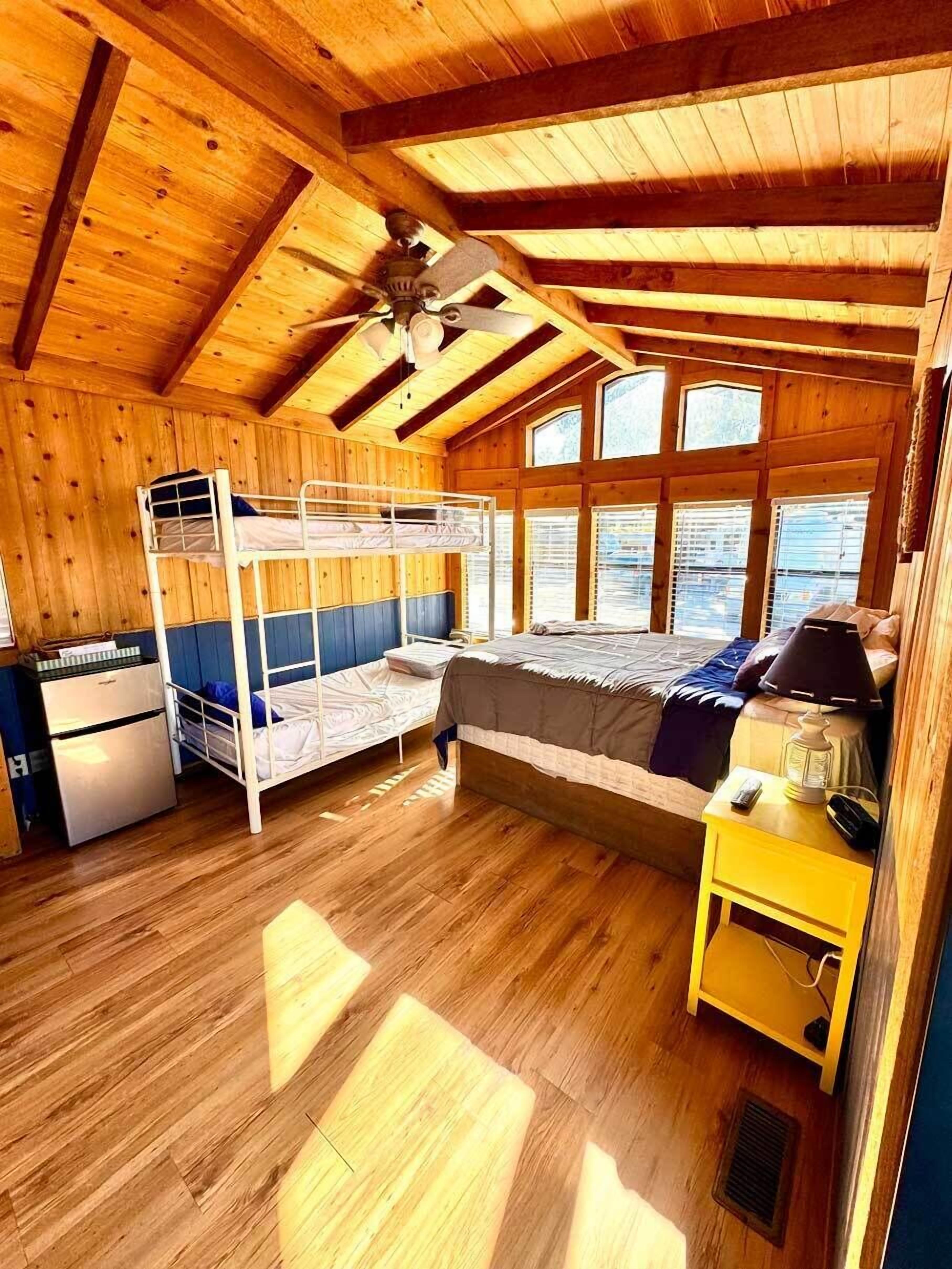 A wooden cabin bedroom with bunk beds, a desk, a refrigerator, and a window with a view.