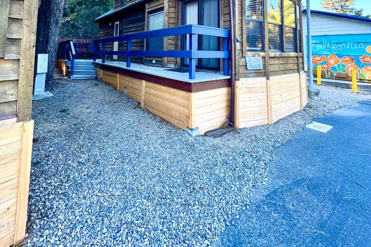 A wooden cabin with a covered porch and a small deck, surrounded by gravel and trees.
