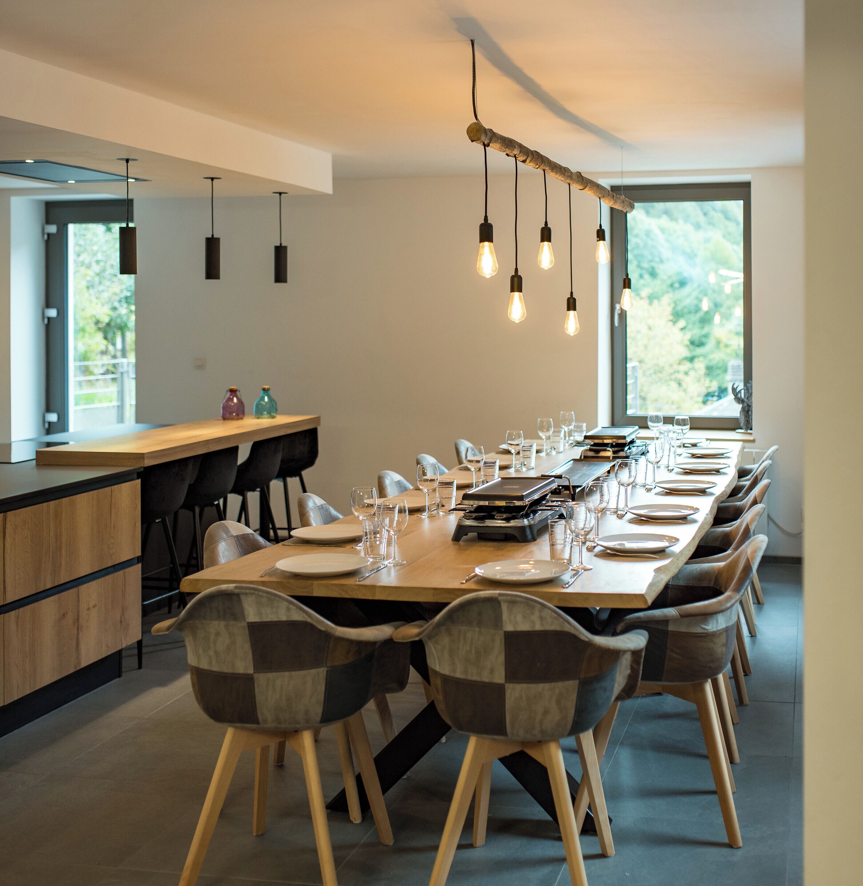 Un espace de restauration moderne, avec une longue table en bois dressée pour un repas, entourée de chaises rembourrées aux pieds en bois.