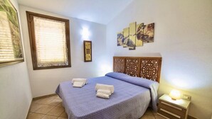 A bedroom with a bed, a nightstand, a wall-mounted artwork, and a window with blinds. - Porto Corallo Two-room apartment 4 beds (Villaputzu)