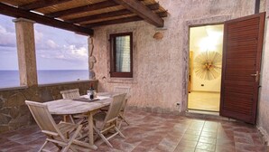 Eine überdachte Terrasse mit Tisch und Stühlen, Blick auf das Meer und einer Tür zu einem Innenraum.