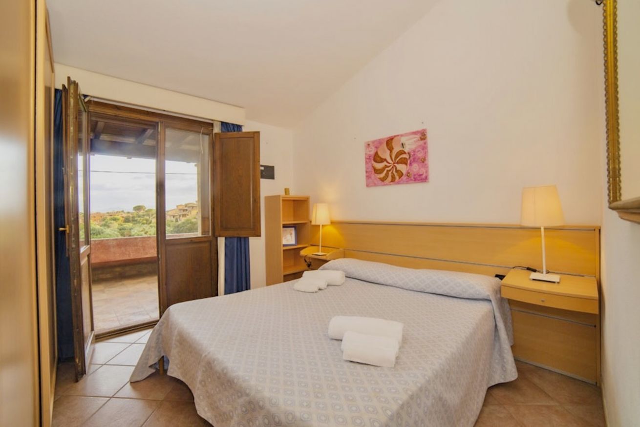 Habitación de hotel con cama, mesita de noche, lámpara, un cuadro y puerta corrediza a un balcón.